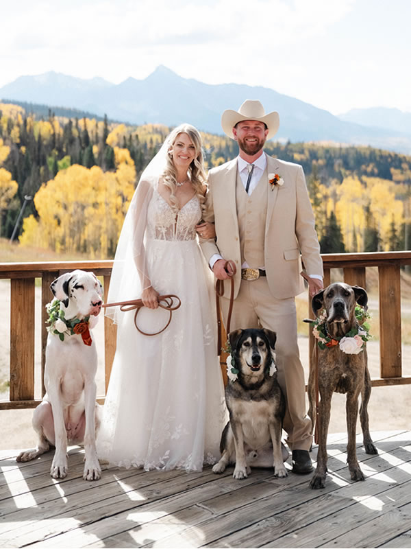 A bride and a groom in a cowboy hat with their three dogs in floral collars on a mountain deck