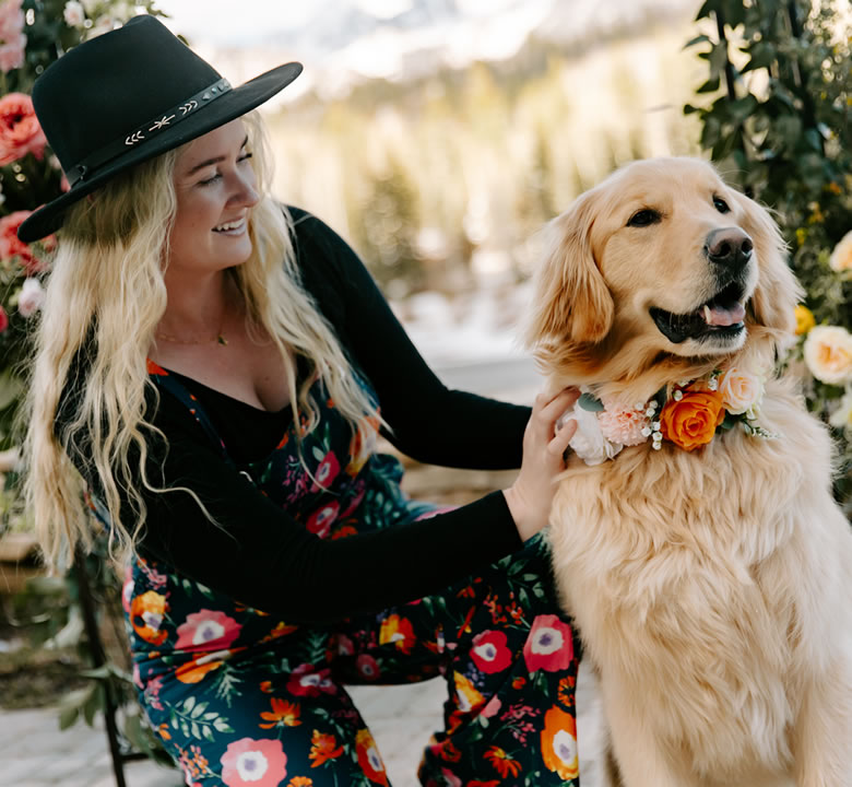 Megan Logsdon in a black hat kneeling beside a golden retriever wearing a floral collar at a wedding