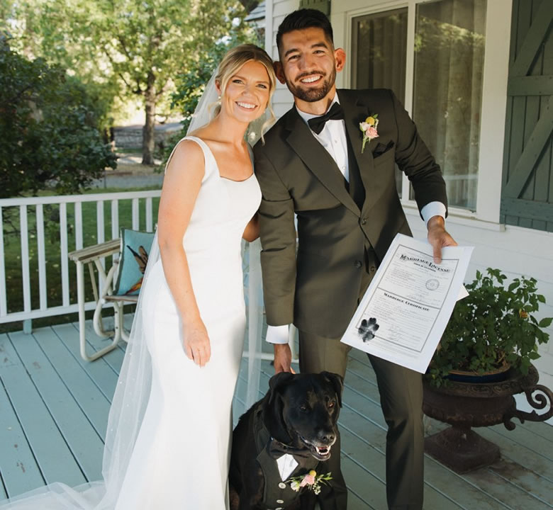 Newlyweds holding their marriage licence on a porch with their black dog in a tuxedo and floral collar